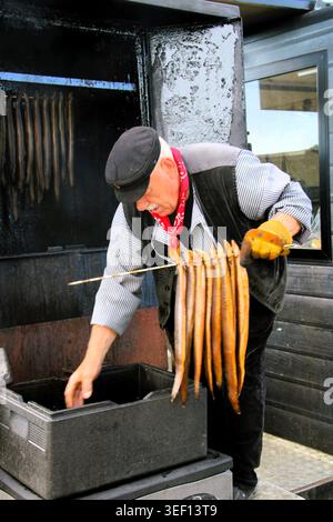 Man leans over smoker, handling fish with care. Fish hang suspended, ready for curing. Steam rises as he checks the batch. Focused, he ensures perfect Stock Photo