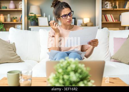 Young woman wearing glasses reviewing documents while using laptop at home, focused on paperwork, online study, remote job, planning tasks and freelan Stock Photo