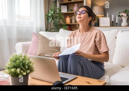 Young woman wearing glasses reviewing documents while using laptop at home, focused on paperwork, online study, remote job, planning tasks and freelan Stock Photo