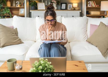 Young woman wearing glasses reviewing documents while using laptop at home, focused on paperwork, online study, remote job, planning tasks and freelan Stock Photo