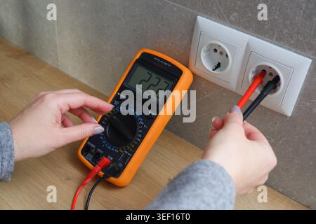 Woman with a multimeter check the voltage of the electrical network. Close-up view of the hands with the appliance near the socket. Stock Photo
