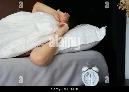 A serene image of a woman resting comfortably in bed, covered with a white blanket and pillow, with a white alarm clock on the bedside table, conveyin Stock Photo