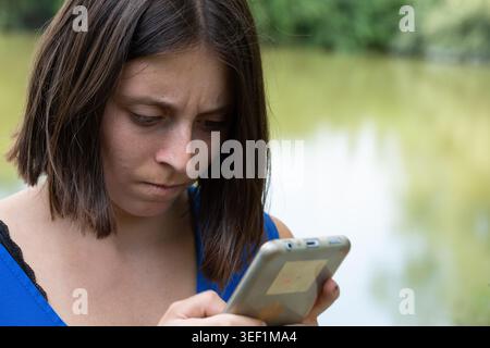 Young woman looking at smartphone with a serious, thoughtful expression by a lake Stock Photo
