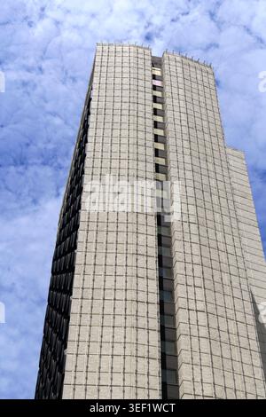 Modern skyscraper with a textured grid facade, casting shadows under a vibrant blue sky filled with white fluffy clouds, representing urban developmen Stock Photo