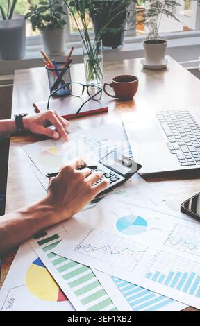 Person working on financial documents with calculator and charts. Hands focus on data analysis on wooden desk. Natural light from window illuminates w Stock Photo