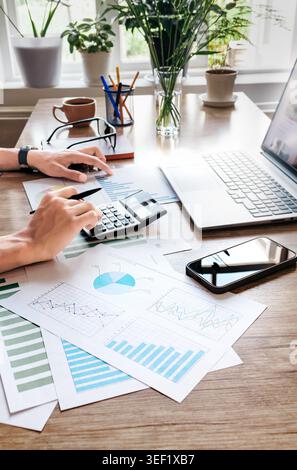Person analyzes financial charts on wooden desk. Hands use calculator and mouse near laptop. Papers show colorful bar and pie graphs. Coffee mug and g Stock Photo