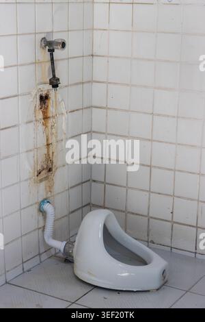 A dirty, broken squat toilet lying on a tiled corner with exposed plumbing, rust stains and water damage. Stock Photo