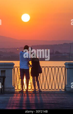 Couple sunset romantic, rear view of a young couple taking a photo of a sunset over the bay in Ortigia, near Syracuse in Sicily. Stock Photo