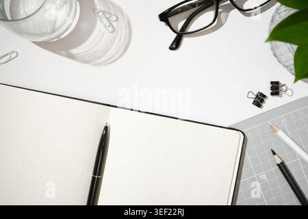 Flat lay of open dotted notebook, black pen, glasses, paper clips, binder clips, pencils, and cutting mat on white desk background. Copyspace Stock Photo