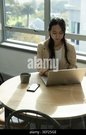 Asian woman wearing beige blazer and necklace, sitting in workspace using laptop, mug, smartphone Stock Photo