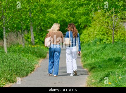 Pair of young women from behind, walking along a country path carrying shoulder bags and talking, on a sunny Spring day in West Sussex, UK. Lifestyle. Stock Photo