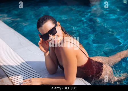 Young woman leaning near pool with radiant wet appearance. Pretty woman enjoying tropical swimming resort during sunny travel holiday. Summer leisure Stock Photo