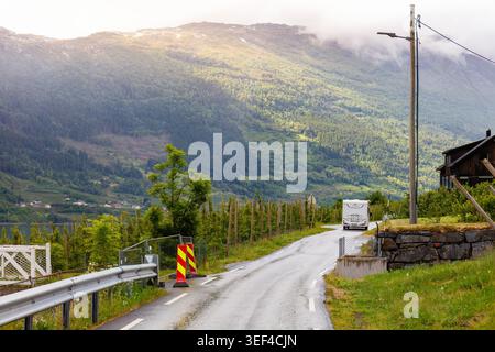 Scenic fjord journey camper vehicle driving along wet mountain road through orchard fields near Hardangerfjord in Lofthus Norway showing slow Nordic t Stock Photo