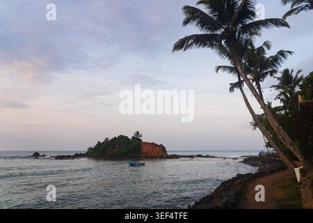 Scenic view of Parrot Rock island in Mirissa, Sri Lanka, seen from the coastline with tall coconut palm trees in the foreground at dusk. Stock Photo