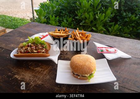 Fast food meal served on a rustic wooden outdoor table, including a sesame seed bun hamburger, a loaded hot dog or sub sandwich Stock Photo