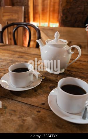 Two white cups of black tea with saucers and a ceramic teapot on a rustic wooden table in a cafe, Sri Lanka. Stock Photo