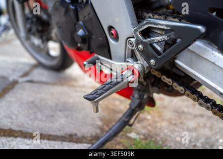 Closeup footpeg and kickstand near chain on parked motorcycle, red frame and pavement textures in frame, Stock Photo