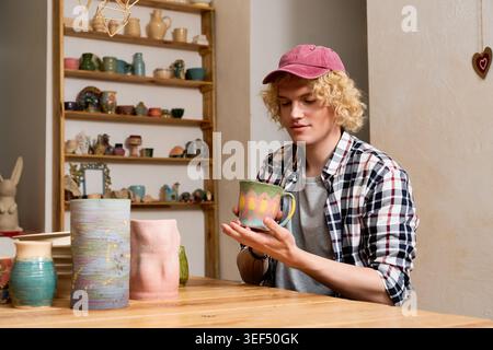 Blonde potter carefully examines his vibrant mug creation in a cozy workshop. Stock Photo