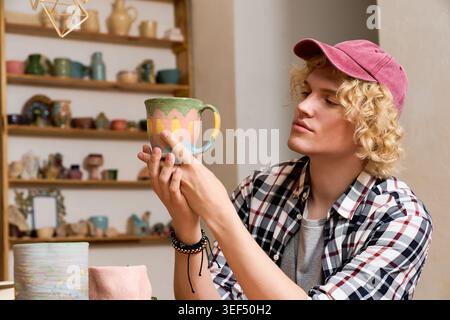 Young man carefully examines a colorful ceramic mug in his pottery workshop, showcasing skill Stock Photo
