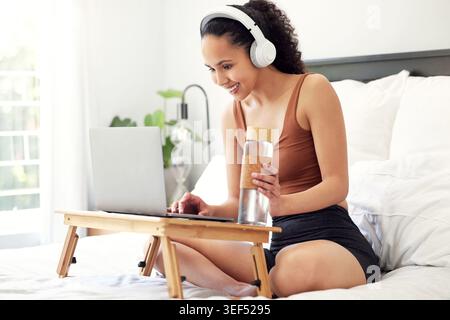 Headphones, laptop and woman on bed in home with listening to music, album or podcast online. Audio tech, water bottle and female person with Stock Photo