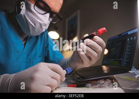 Medical professional examining a blood test tube during laboratory analysis with a laptop Stock Photo