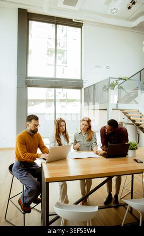 Group of four people collaborates on a project in a well-lit office. They use laptops and discuss ideas while seated around a wooden table. Sunlight c Stock Photo