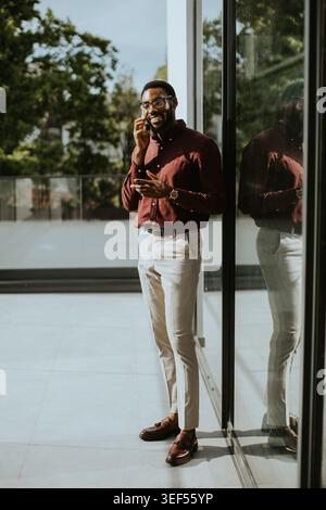 A man stands by a glass door in a modern building. He is talking on his phone while checking his watch. The scene shows greenery outside the window an Stock Photo