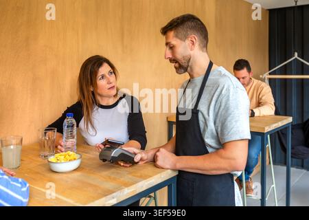 Female customer making contactless payment using smartphone in cafe while talking with waiter Stock Photo