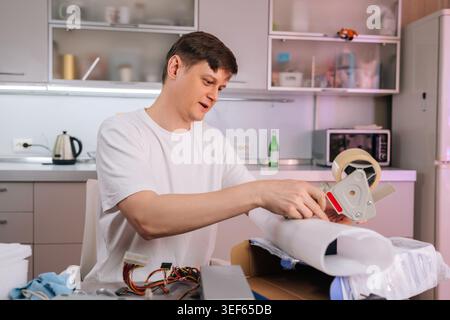 Young caucasian man packs computer components into shipping box at home. He using tape dispenser and wrapping material. Concept for ecommerce, small Stock Photo