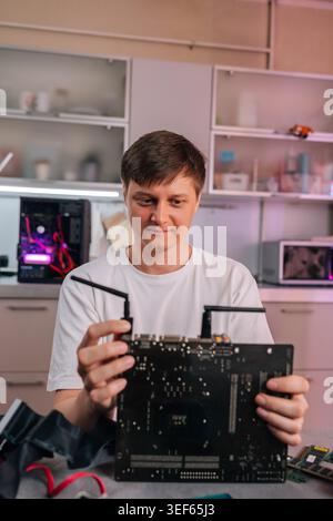 Focused man in white shirt examining computer motherboard. Technician works on hardware assembly or maintenance in home workshop with purple light in Stock Photo