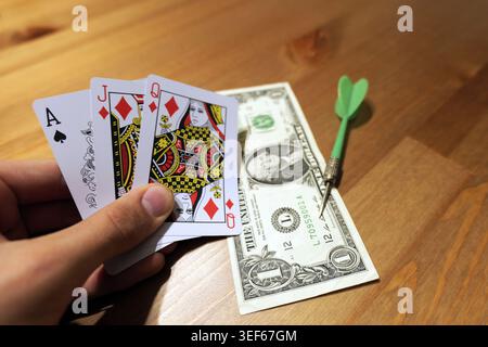 Hand holding an ace, jack, and queen of diamonds while a dollar bill and a green dart are resting on a wooden table, representing risk, strategy, and Stock Photo