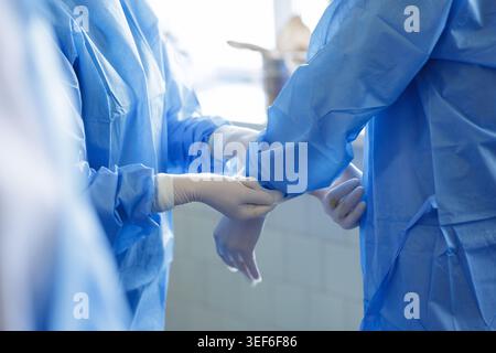 Two medical professionals assist each other in putting on surgical gowns and gloves in a bright operating room, showcasing teamwork and preparation fo Stock Photo