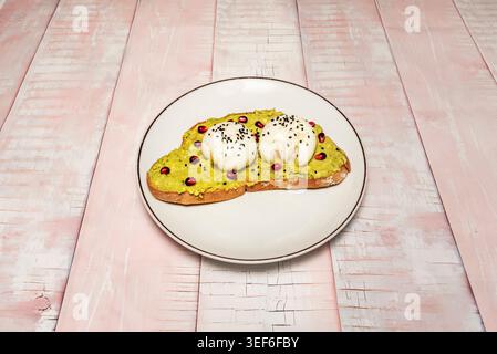 Smashed avocado toast topped with two burrata balls, sesame seeds and pomegranate sits on a white plate over pink wood. Stock Photo