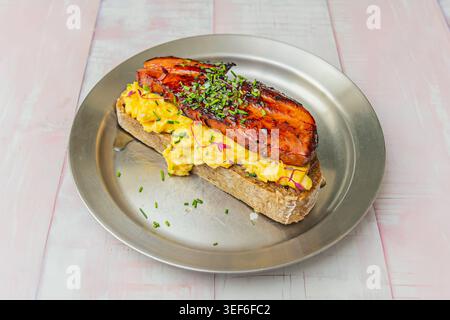 Open toast topped with creamy scrambled eggs, crispy glazed bacon and chopped chives on a metal plate over whitewashed wood. Stock Photo