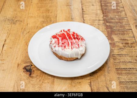 A glazed round pastry with red crumb topping and white drizzle sits on a white plate over a rustic wooden table. Stock Photo
