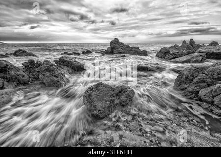 A Closeup Of Water Flowing Over Rocks With The Sun Breaking Through Clouds On The Ocean Horizon Black And White Stock Photo