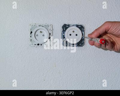 Installing a new electrical wall socket during renovation work indoors. Hand using voltage tester near electrical installation on white wall. Stock Photo