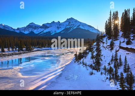 Frozen River Winter Forest and Sunburst Over Mountain Landscape Stock Photo