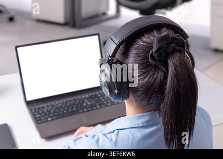 Woman wearing headphones while working on laptop mockup in modern office, representing online learning, remote meetings and digital communication conc Stock Photo