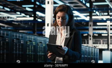 Engineer walking in data center, monitoring industrial automation systems using tablet. Server room IT expert using device to oversee network equipment performance Stock Photo