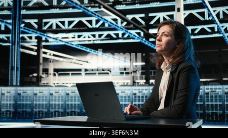 Data center software developer optimizing server load balancing using laptop. Woman in server farm at desk adjusting system parameters on notebook to improve efficiency Stock Photo