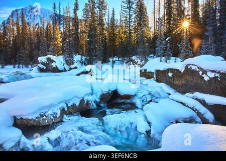 Frozen River and Snow Covered Natural Bridge with Sunburst Through Winter Forest Stock Photo