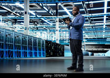 Data center programmer reviewing network configuration with tablet, running predictive maintenance. African american man in server farm scanning hardware logs, tracking computer bugs using notebook Stock Photo