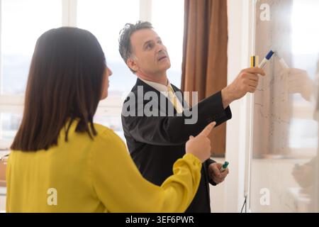 A male professor in a suit stands at a whiteboard, pointing at complex mathematical physics and structural engineering equations while two female stud Stock Photo