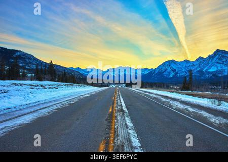 Winter Sunrise Road Leading to Snowy Mountain Range and Pine Forest Stock Photo