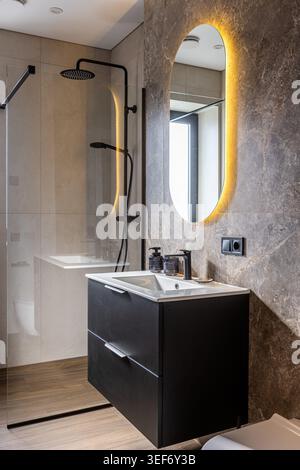 A contemporary bathroom design featuring a dark floating vanity cabinet, a white sink, black fixtures, and an oval mirror with warm LED backlighting a Stock Photo