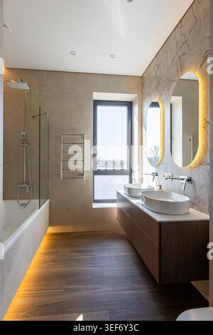 A spacious modern bathroom featuring a wooden double floating vanity with white vessel sinks, oval backlit mirrors, a bathtub with a glass shower scre Stock Photo