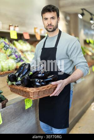 Careful middle aged salesman placing eggplants on counter in large grocery store Stock Photo