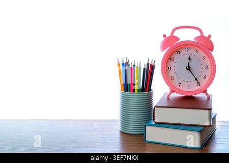 A pink alarm clock on stacked books beside a cup of colored pencils on a wooden desk. Study time, morning routines, and school scheduling concepts. Stock Photo