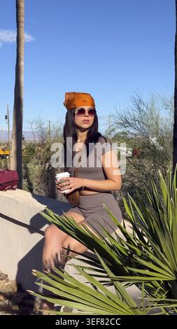Young woman sitting on a wall taking a break during her journey, wearing a headscarf and sunglasses while holding a coffee cup in a desert landscape u Stock Photo